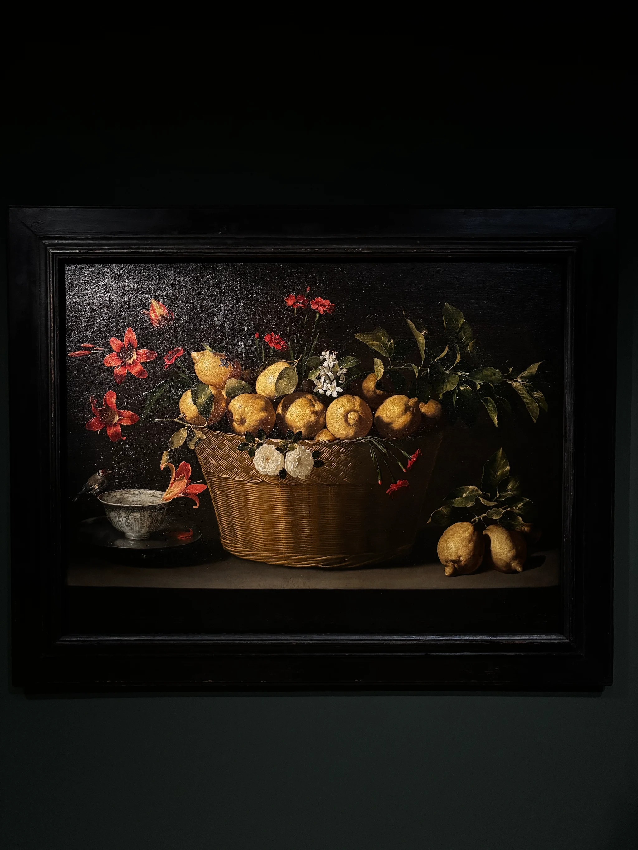 A still life by Juan de Zurbarán in a dark frame: a wicker basket heaped with lemons and their leaves, red carnations and lilies, white roses at the basket rim and orange blossom above, a goldfinch perched on a blue-and-white porcelain cup on a silver plate at left, and more lemons on a leafy branch on the table at right, all against a black ground.