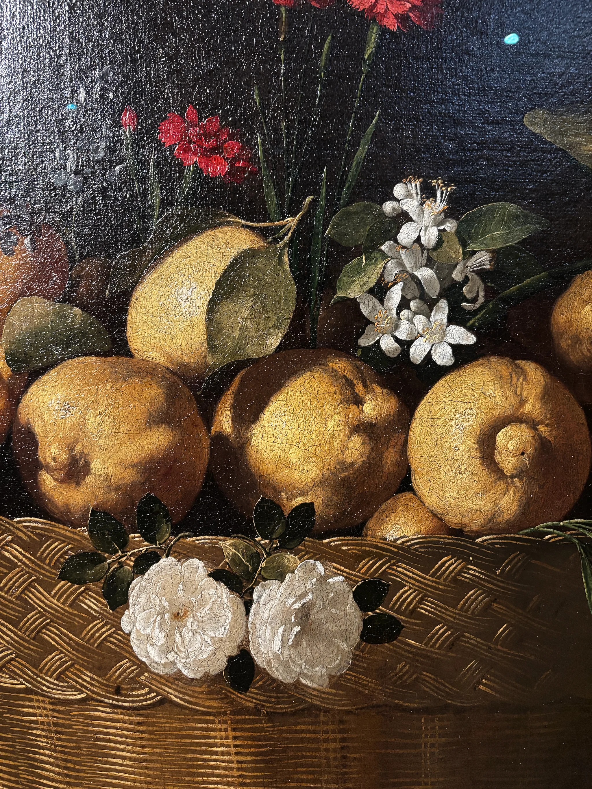 A close view of the same still life: lemons with craquelure running across the paint, white roses and dark leaves resting on the woven rim of the basket, orange blossom and a red carnation above, the weave of the wicker rendered stroke by stroke.