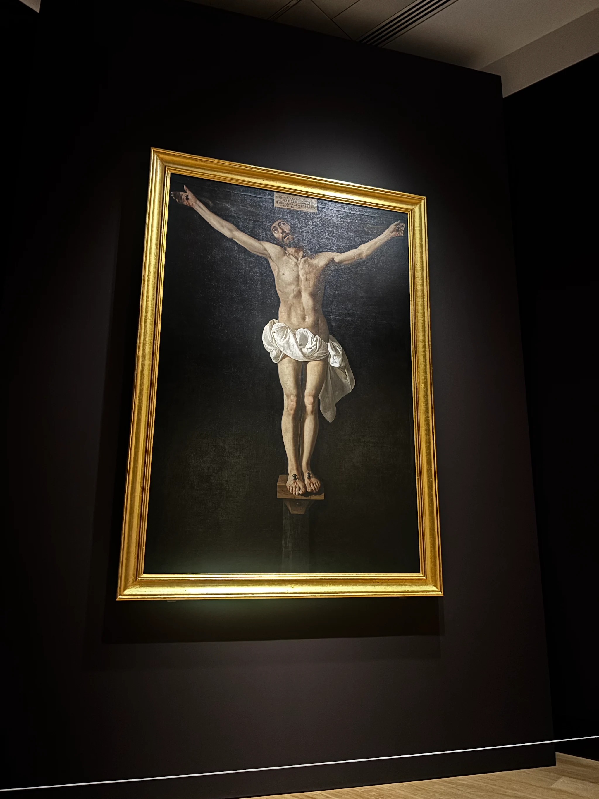 A large crucifixion in a gold frame on a darkened gallery wall, lit by a single spotlight. Christ, alive and looking upward, hangs against a black ground with a white loincloth, a small cartellino nailed above the cross; the dark wall around the frame falls away into shadow.
