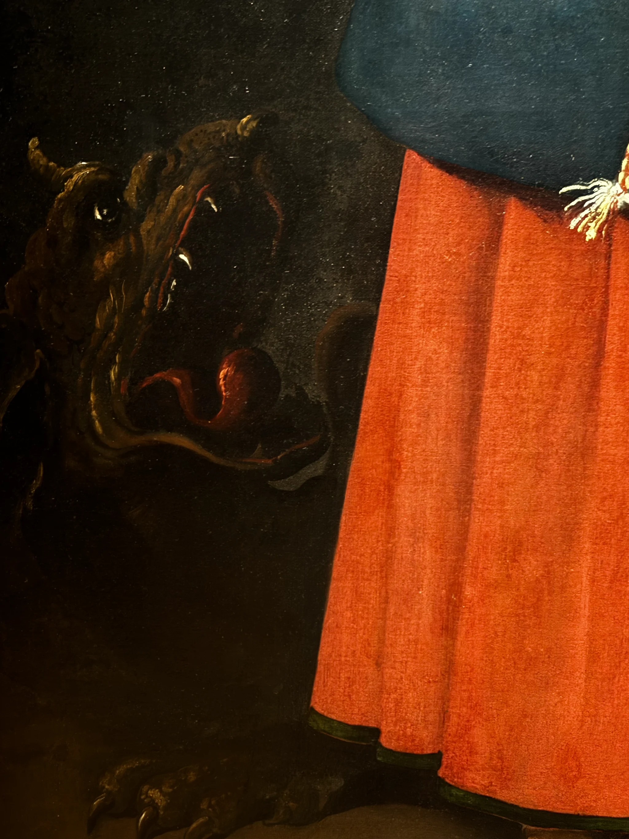 A close view of Saint Margaret of Antioch: the dark horned head of the dragon, jaws open around a red tongue, nearly swallowed by the black ground, beside the saint’s red skirt and blue cloak at right.