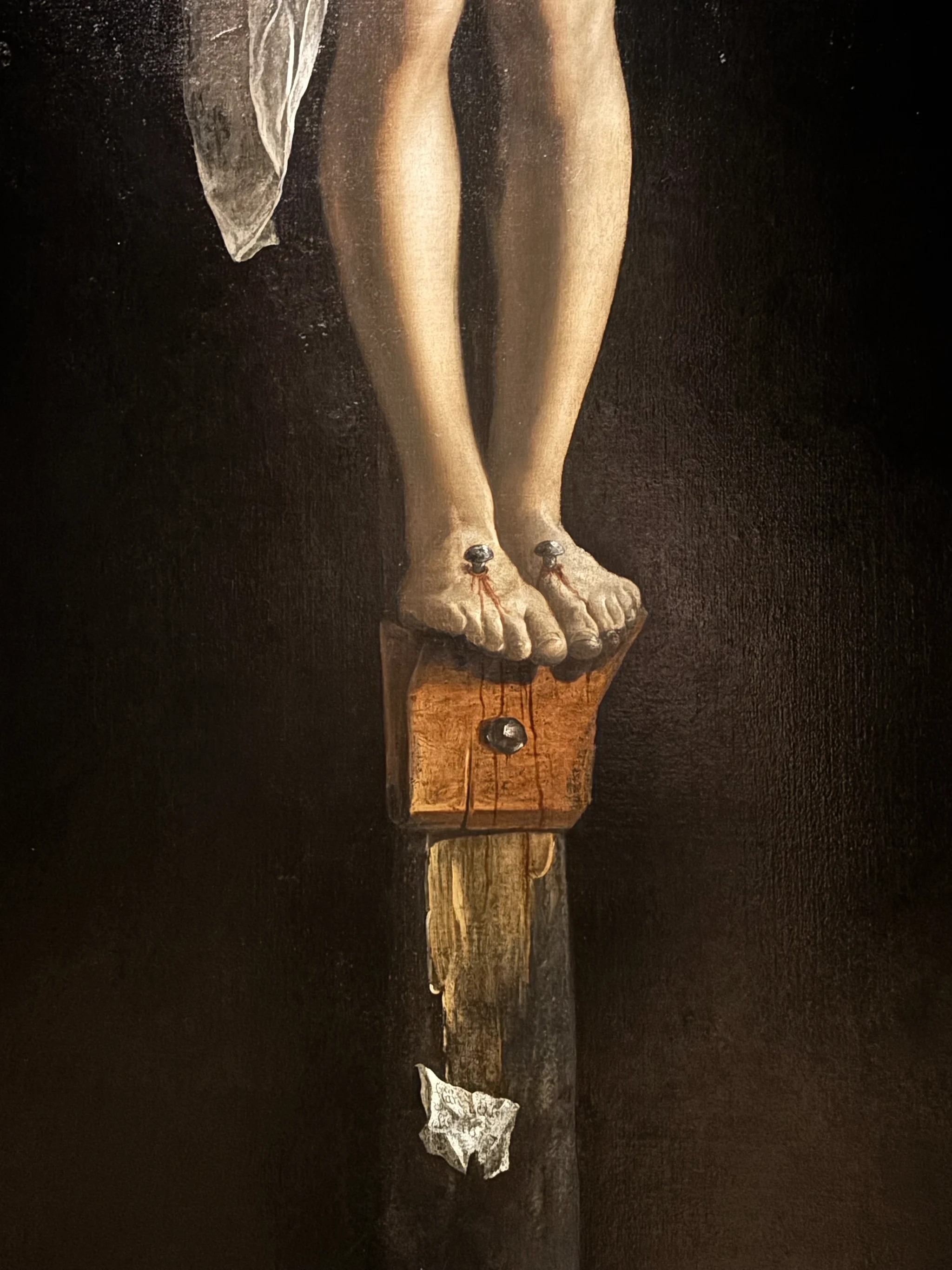 A close view of the crucifixion: two feet nailed side by side to a small wooden block, thin runs of blood from each nail, and further down the post a painted scrap of paper curling away from the wood.