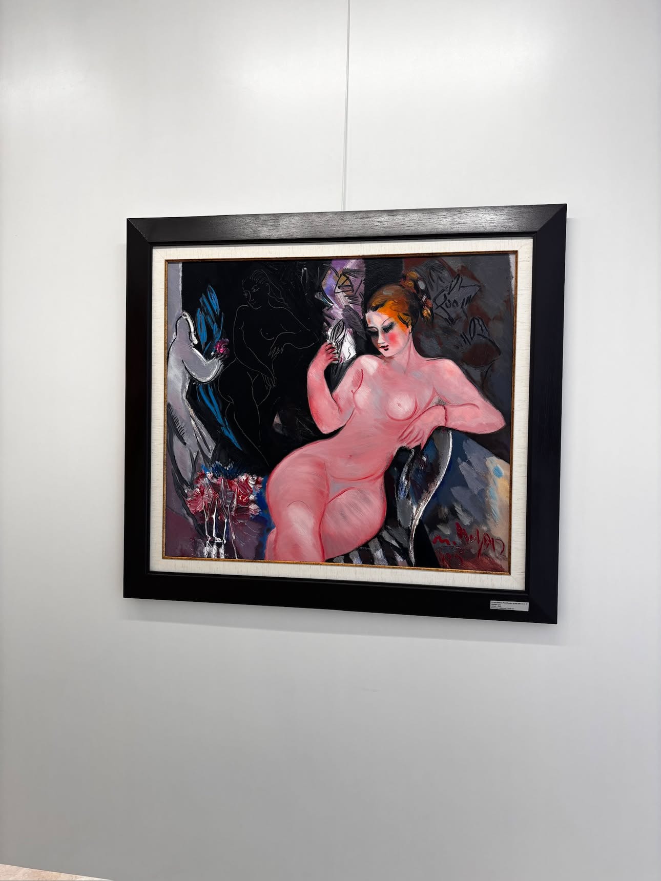 A painting of a pink-toned nude woman reclining and holding a small mirror, a dark cloaked figure faint behind her, in a black frame on a white gallery wall.