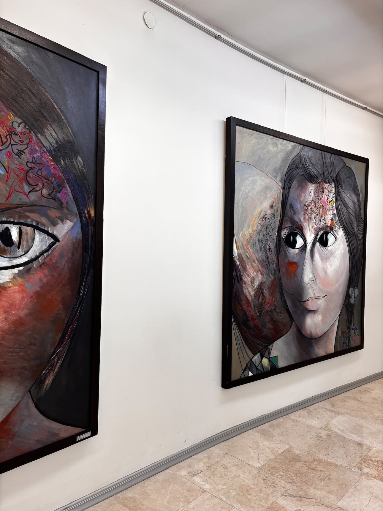 Two large square paintings of close-cropped female faces hung side by side, rendered in dark reds, greys and black with wide, dark eyes.
