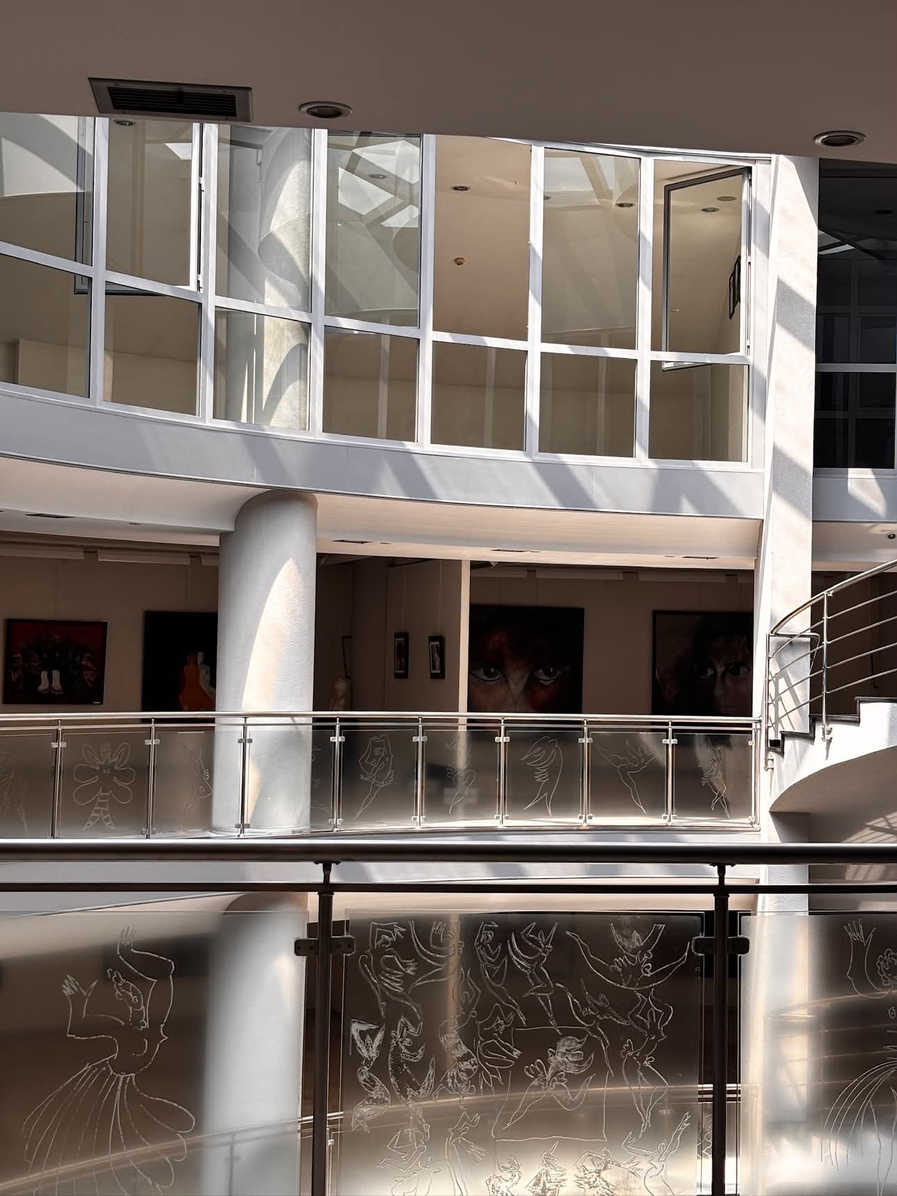 The multi-storey glass and marble atrium interior of the Mustafa Ayaz Museum, paintings visible on the upper-floor walls, and the balustrade glass etched with dancing figures.