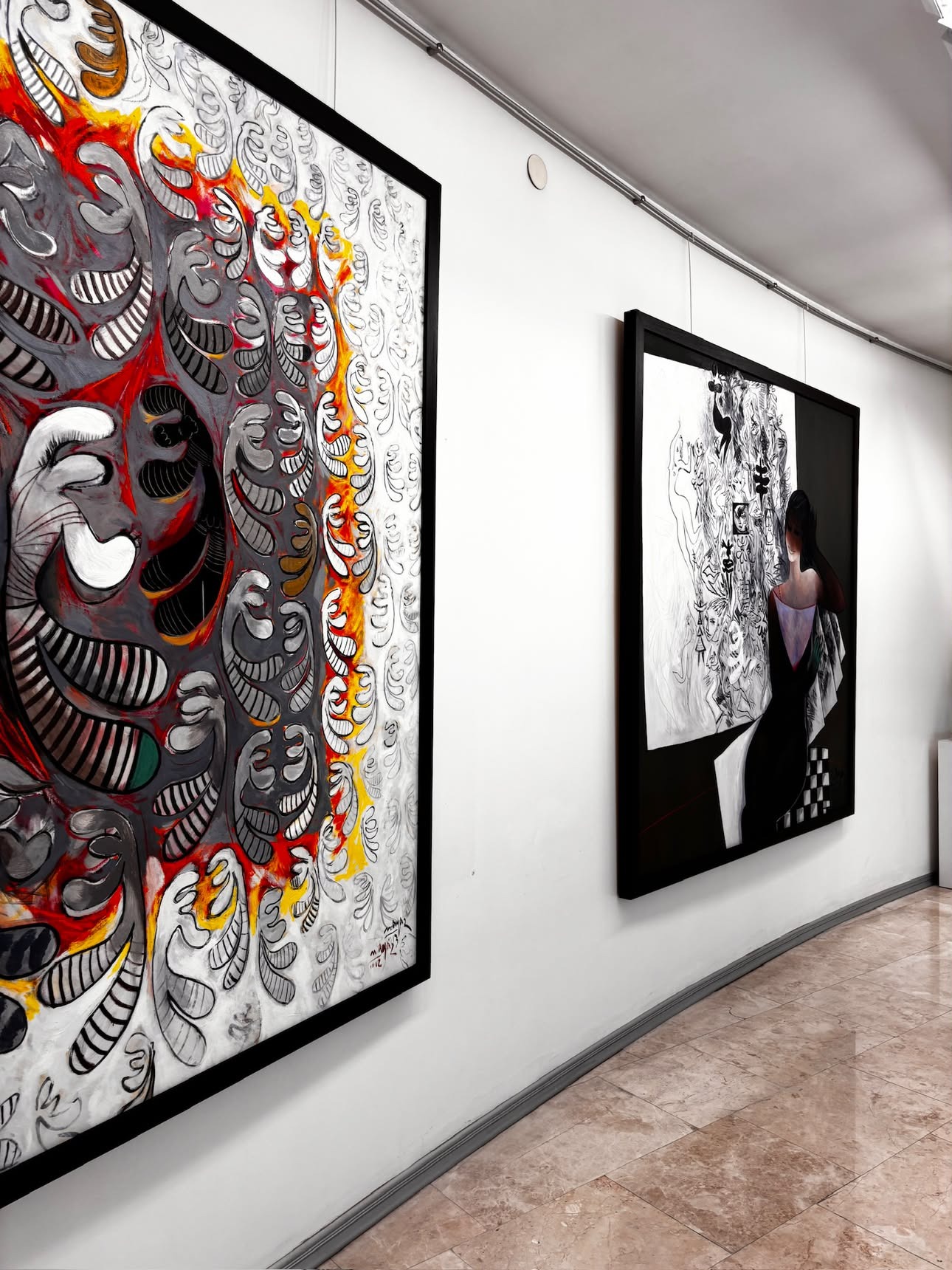 A large painting dense with red, white, grey and black swirling motifs, some resolving into grinning faces, on the left wall, beside a painting of a woman in a pale dress seated against a wall of small sketched black-and-white faces on the right.