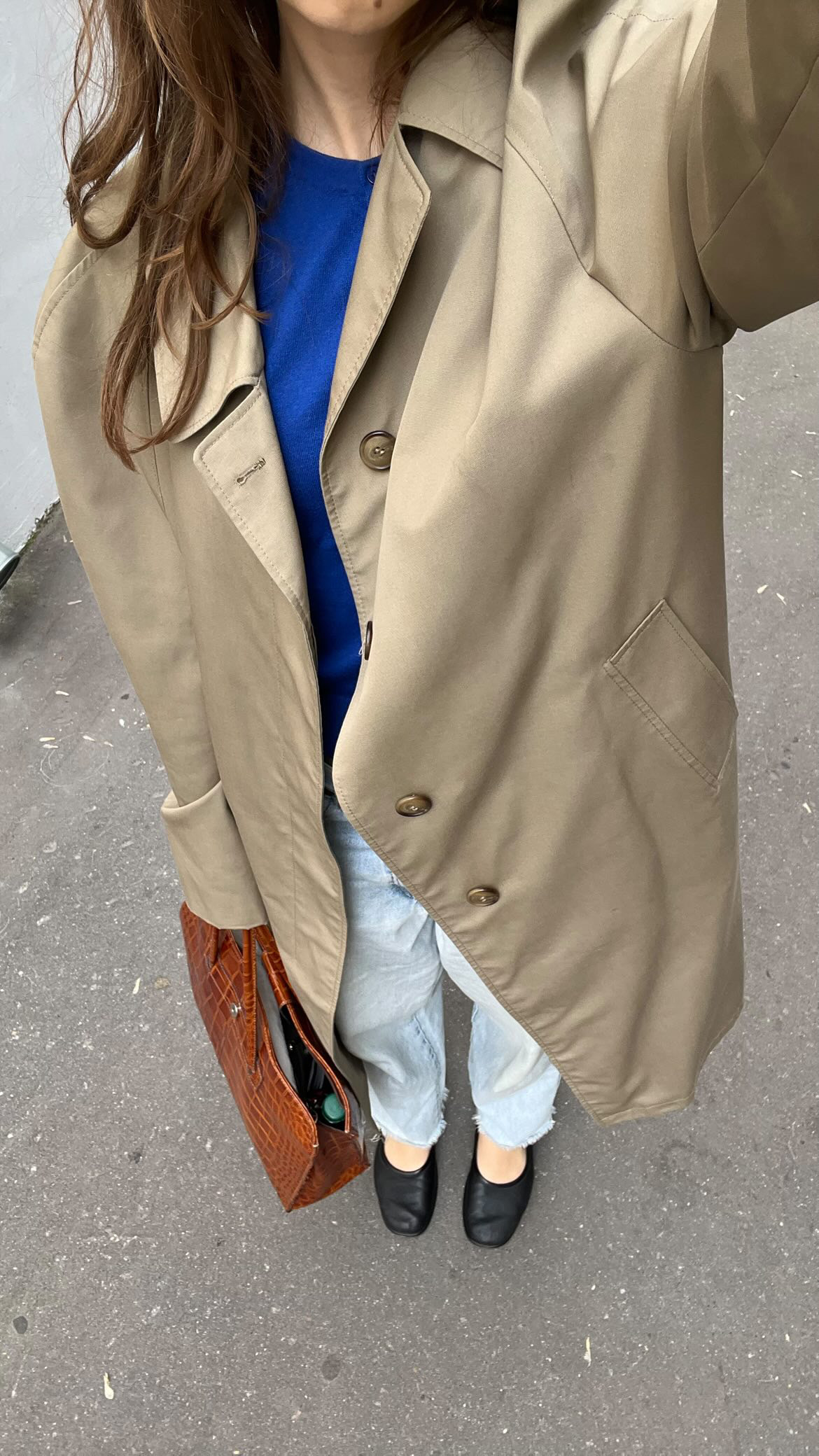 A person photographed from above, from the shoulders down, wearing an oversized stone-coloured trench coat open over a cobalt blue knit, pale blue jeans and black leather flats, holding a tan crocodile-effect bag, standing on grey tarmac.