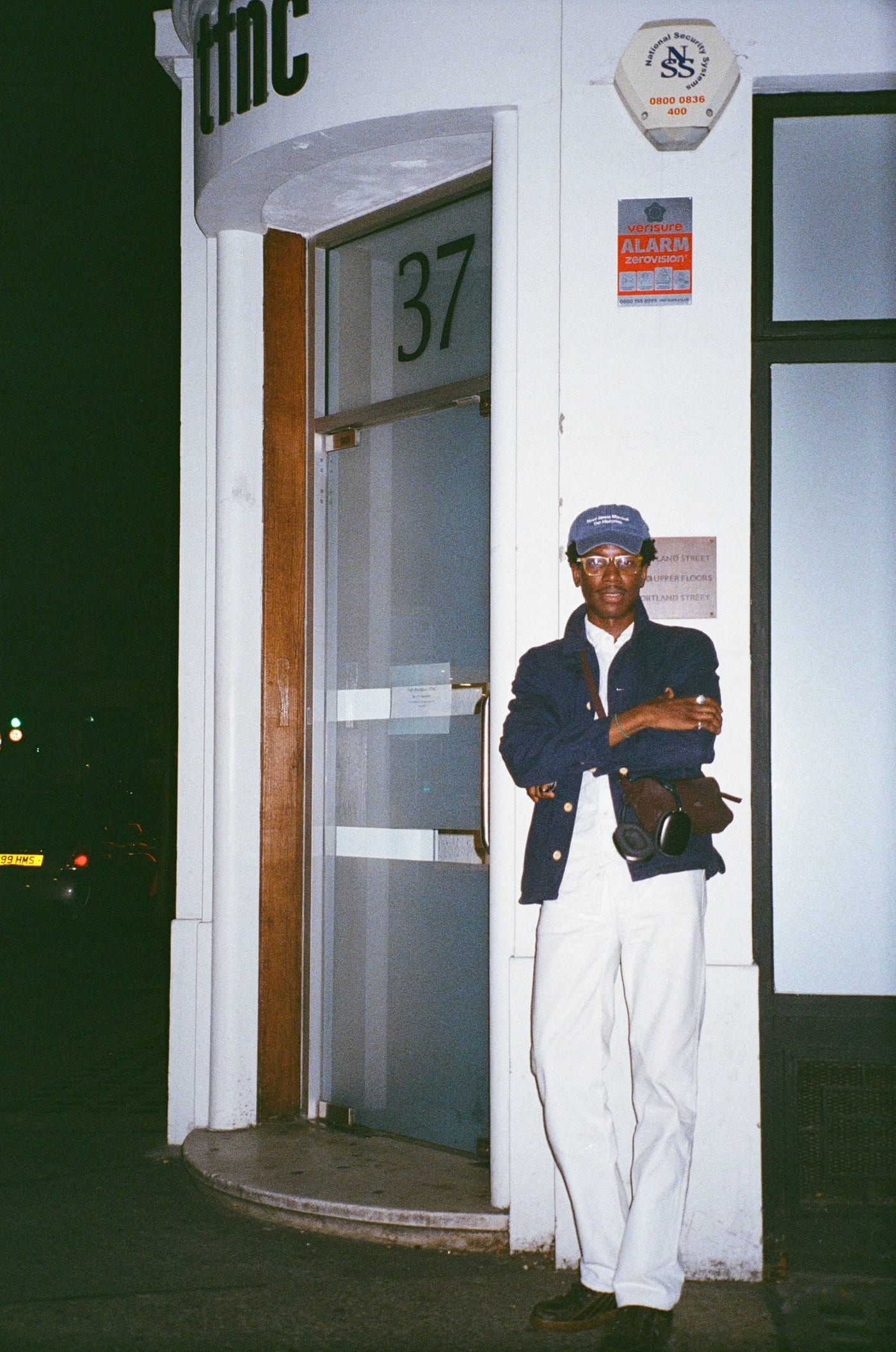 A man standing in the doorway of a white-painted corner building numbered 37, at night, arms folded. He wears a navy worker jacket with pale buttons open over a white shirt, white trousers and black leather shoes, a blue cap, amber-framed glasses, and a brown leather cross-body bag with black over-ear headphones hooked to it.