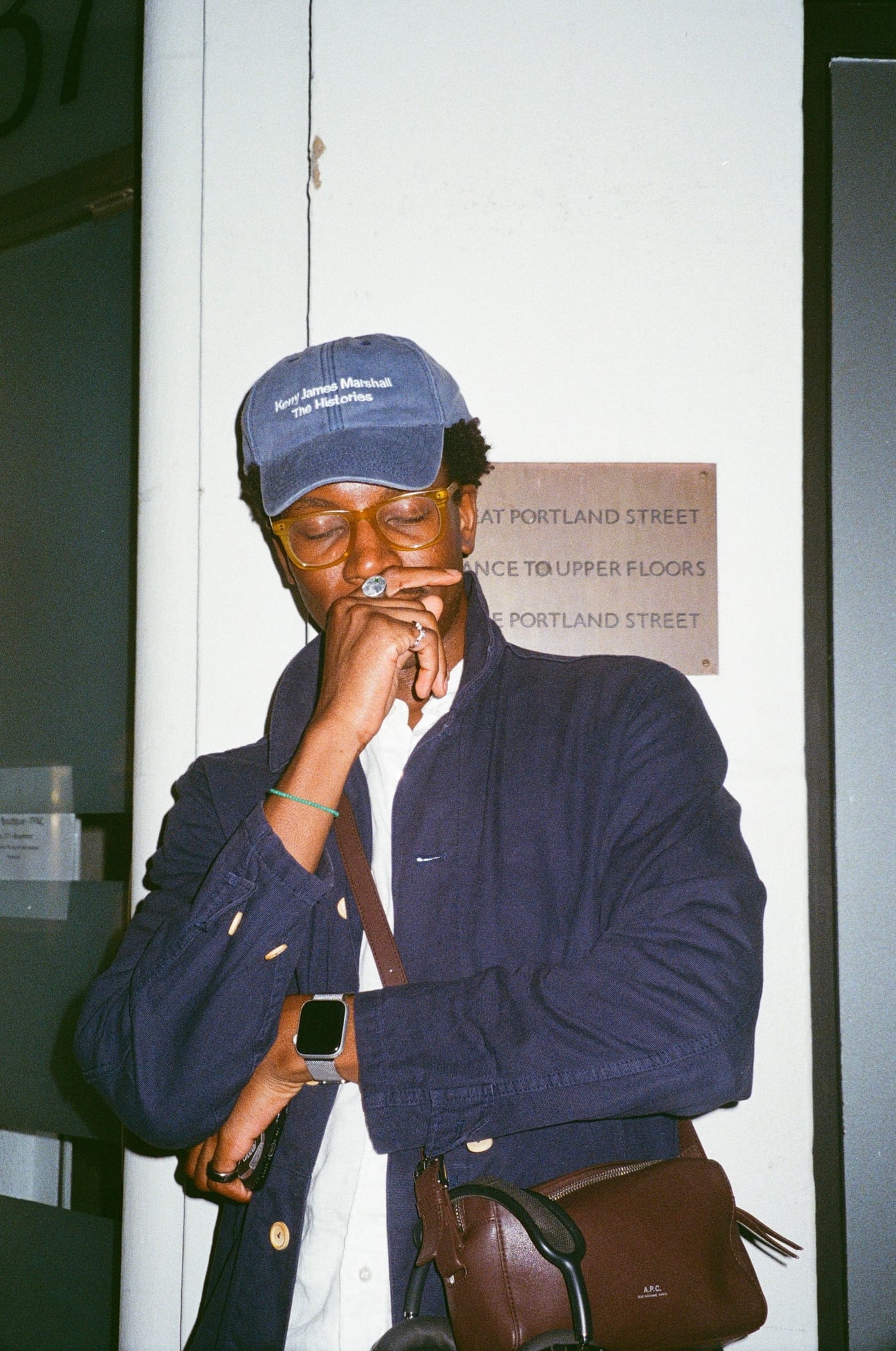 A closer frame of the same man, head lowered and one hand raised to his mouth, several silver rings on his fingers. His blue cap reads Kerry James Marshall, The Histories. A brass plaque behind him reads Great Portland Street, entrance to upper floors.