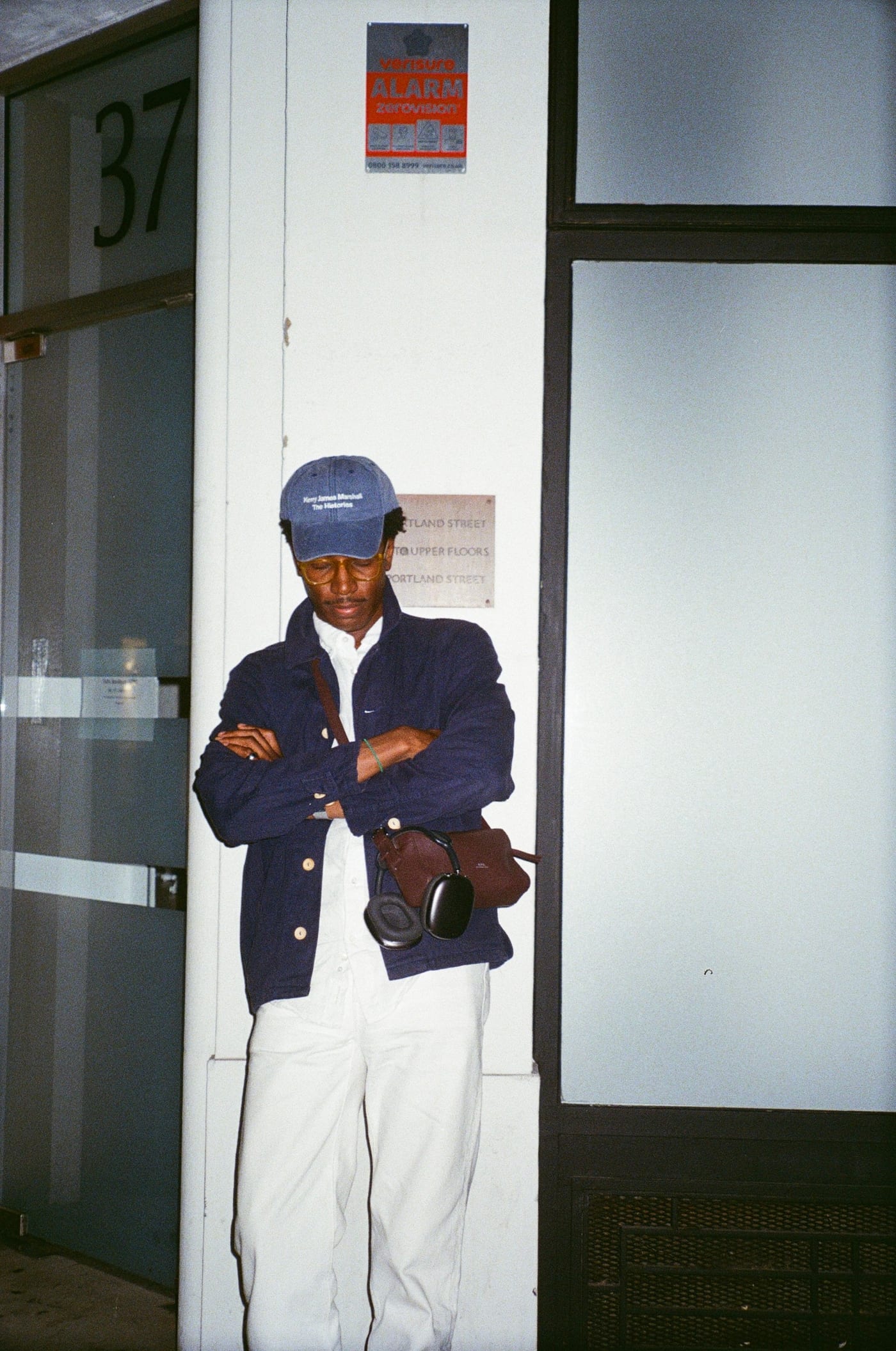 The same man leaning against a white pillar at night, head down and arms folded, in the navy worker jacket, white trousers and black shoes, the brown bag and headphones at his hip. The door number 37 is visible to his left.