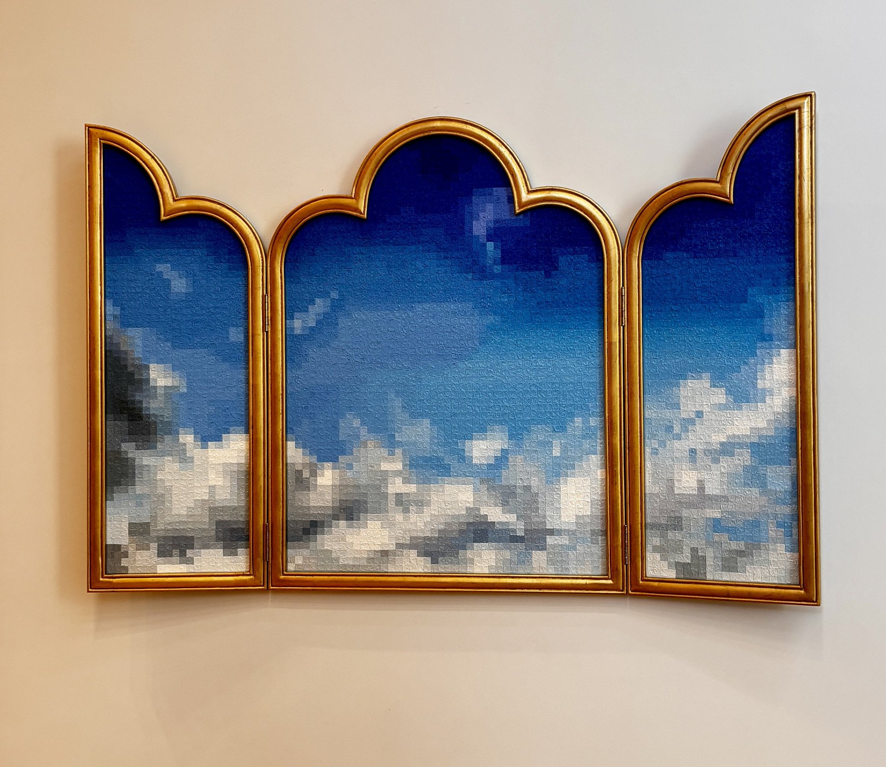 A three-panel painting in an arched gold frame on a pale wall, showing a deep blue sky above banked white and grey cloud, the whole image rendered in visible square pixels.