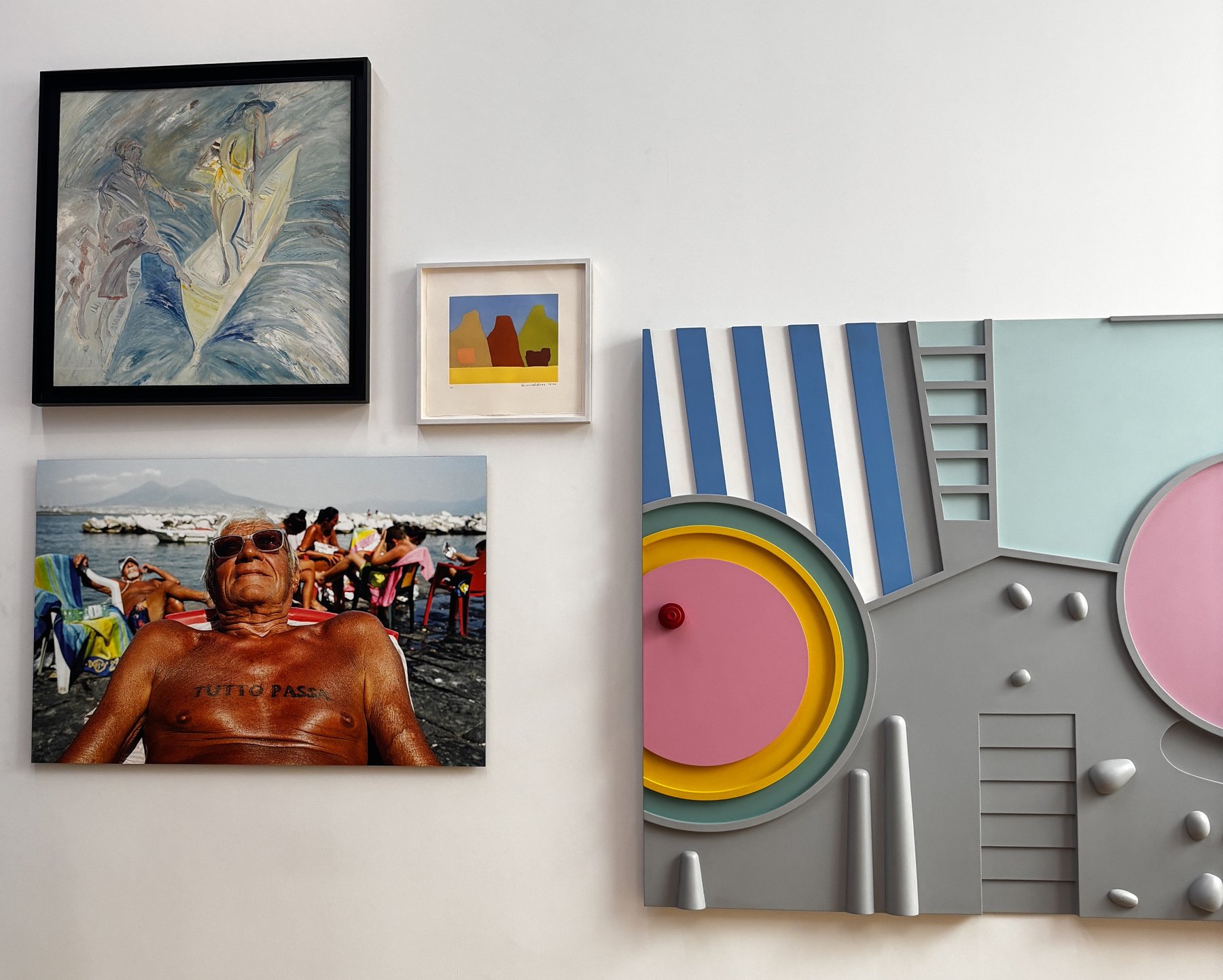 A white gallery wall hung with four works: at upper left a framed painting of pale figures against blue and white; at centre a small framed print of orange and yellow mountain forms; at lower left a photograph of a tanned older man sunbathing; and at right a large relief panel in grey, pink, blue and yellow carrying ladders, stripes and raised dots.