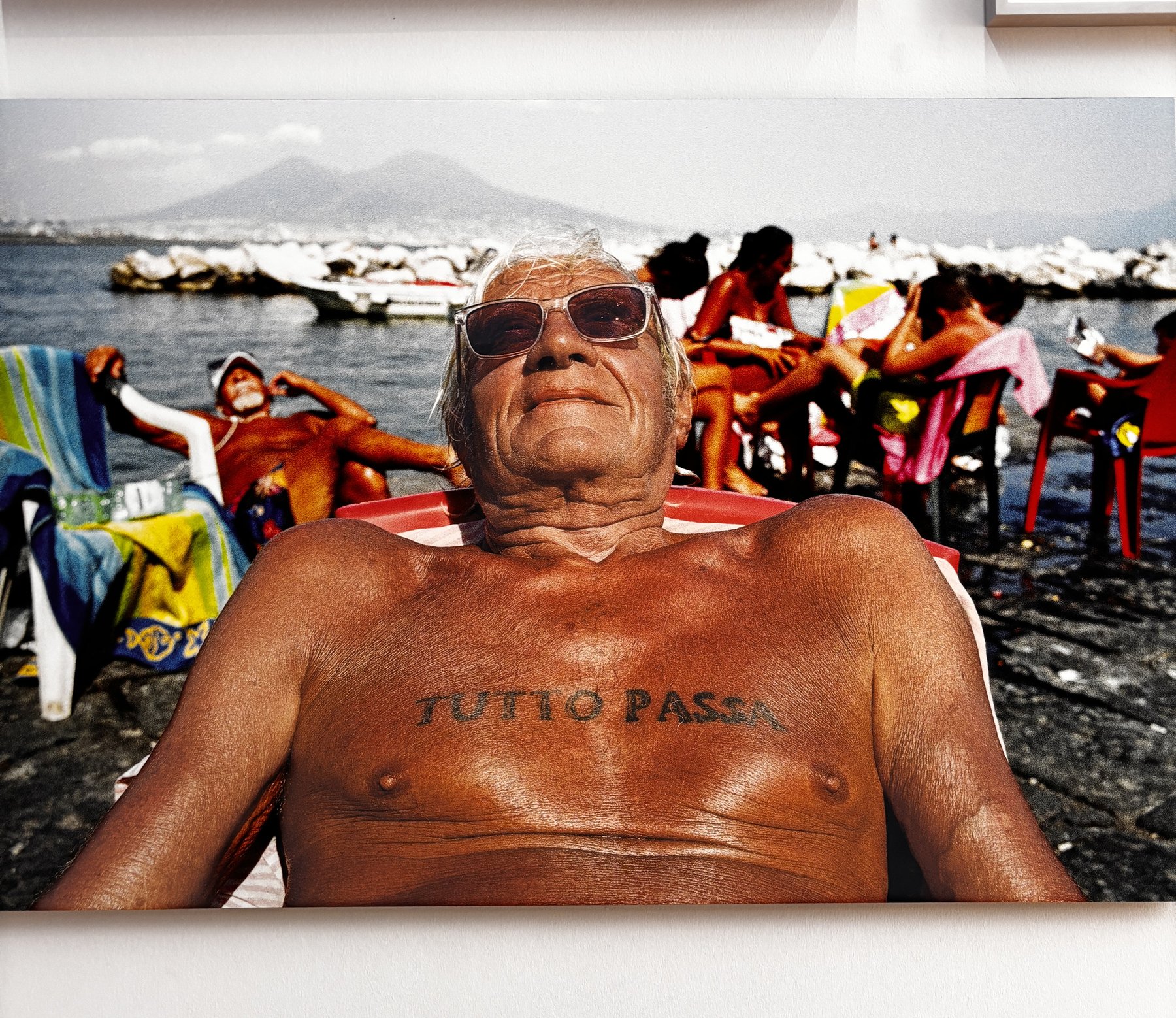 A large photograph on a gallery wall showing a tanned older man in sunglasses reclining in a red and white deckchair on a stony shore, the words TUTTO PASSA tattooed across his chest, with other bathers, plastic chairs and a mountainous coastline behind him.