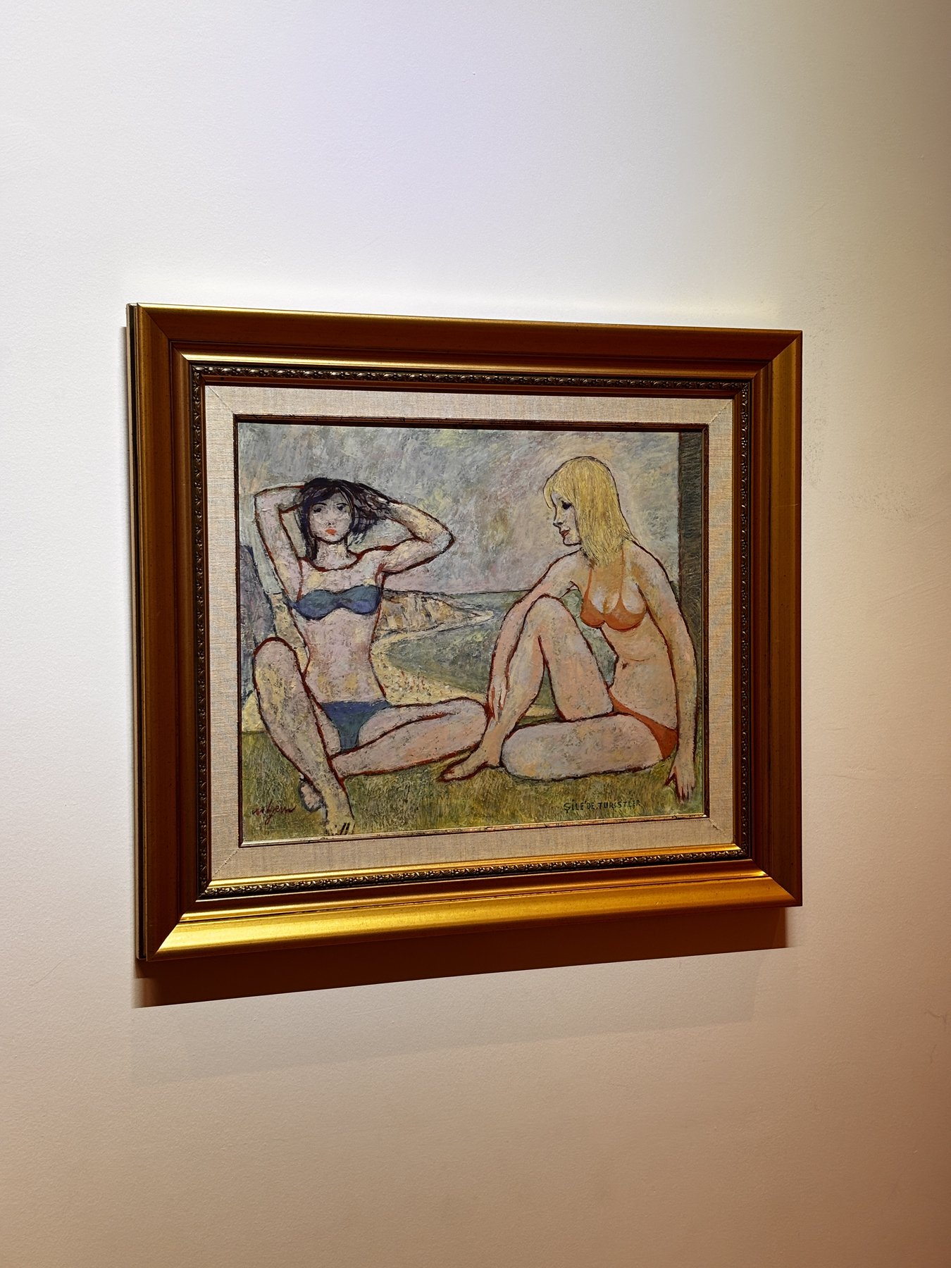 A gold-framed painting of two women resting on grass beside a curving shoreline, one dark-haired in a blue bikini with her arms raised behind her head, the other blonde and bare-chested, seated and turned toward her, the sea and a headland behind them.