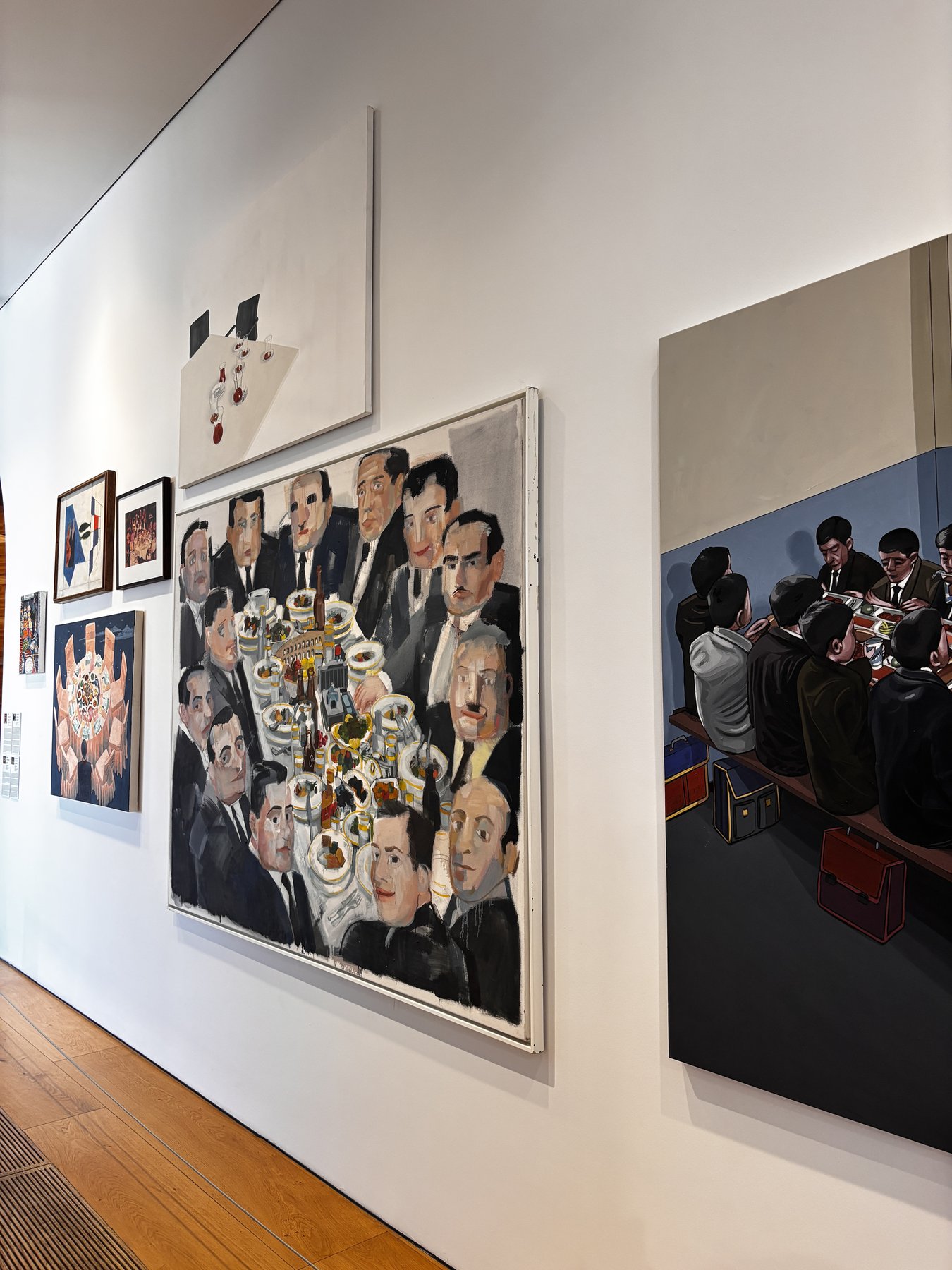 Two large paintings on a white wall. On the left, a crowd of anonymous suited men gathered around a table crowded with white architectural models and vessels. On the right, rows of identically dressed students seen from behind at a long table, briefcases stacked beneath it. Smaller framed works hang further to the left.