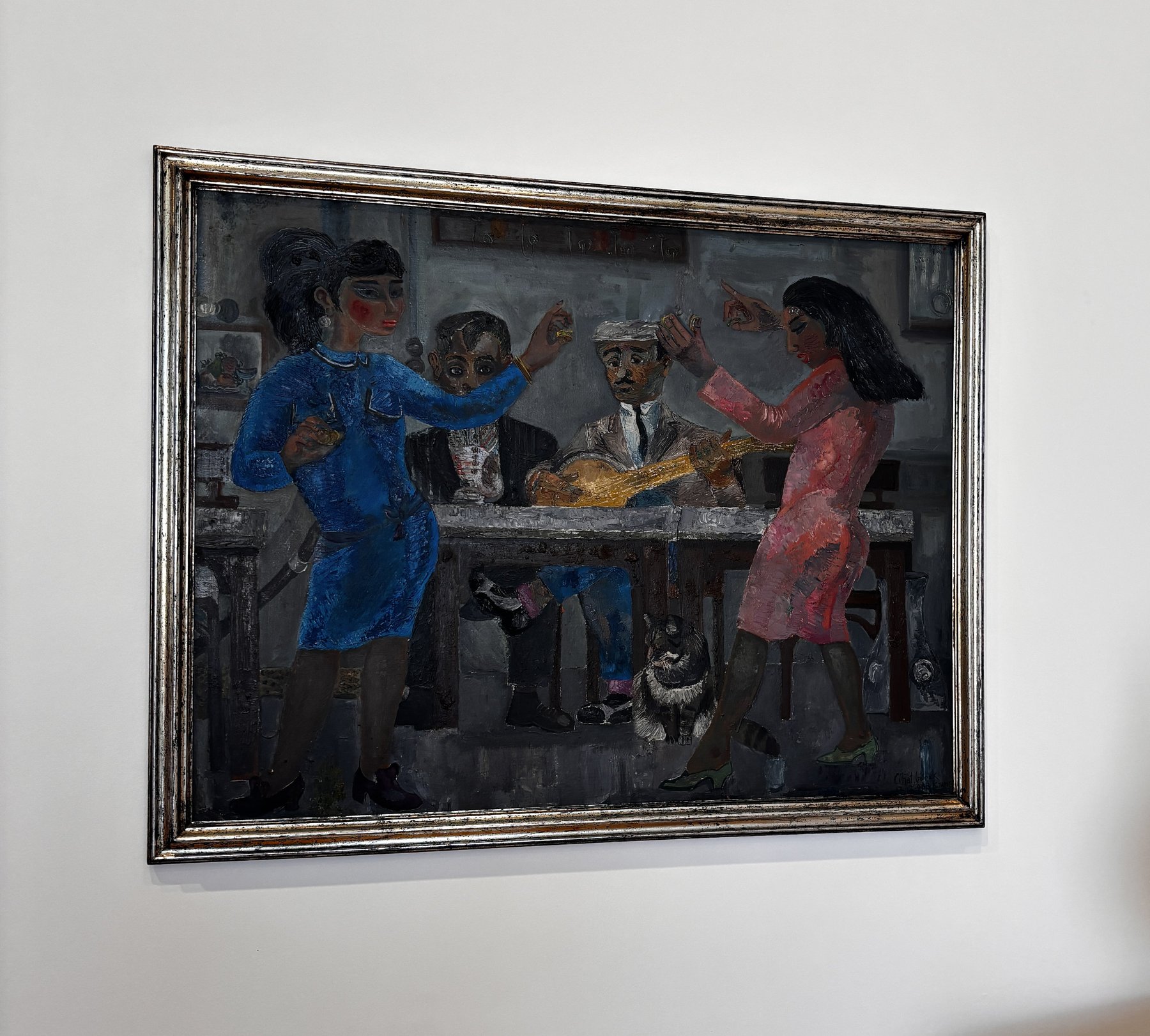 A silver-framed painting of a dim tavern. A woman in blue and a woman in pink dance with raised hands on either side of a table, a seated man plays a round-bodied cümbüş, another man sits behind the table, and a cat sits on the floor beneath them.