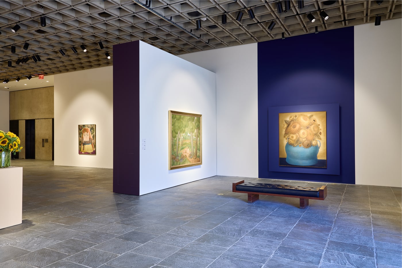 A deep blue wall at right holding the large sunflowers-in-a-blue-jug canvas, a green wooded landscape on white in the centre, a girl in a doorway at far left, and a leather bench.