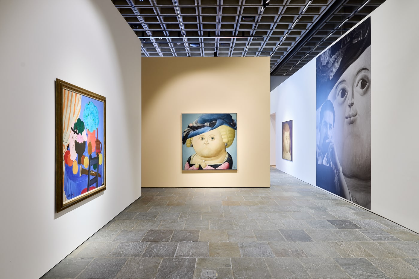 A gold-framed still life with a striped curtain, blue roses and fruit at left, the blue-hatted woman centred on a tan wall, and a tall black-and-white mural pairing a photograph of a man with an enlarged painted face at right.