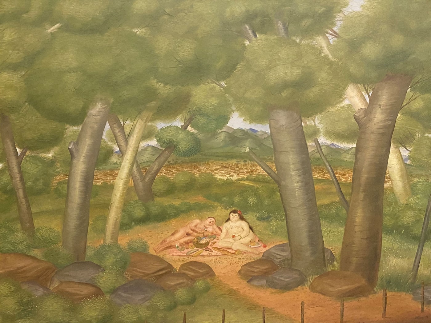 A wooded landscape in soft greens, the upper half filled by rounded canopies on tall pale trunks. In a clearing at the centre a man reclines on his side and a woman sits upright beside him, both nude and heavily rounded, on a pink cloth spread over reddish earth. Between them a wicker basket, loaves, fruit and a small bottle; she holds a red fruit and a plate rests at her side. Beyond the clearing green fields give way to a long band of ochre rooftops and then to blue-green mountains under a pale sky. Grey and brown boulders and a thin post fence close the foreground.