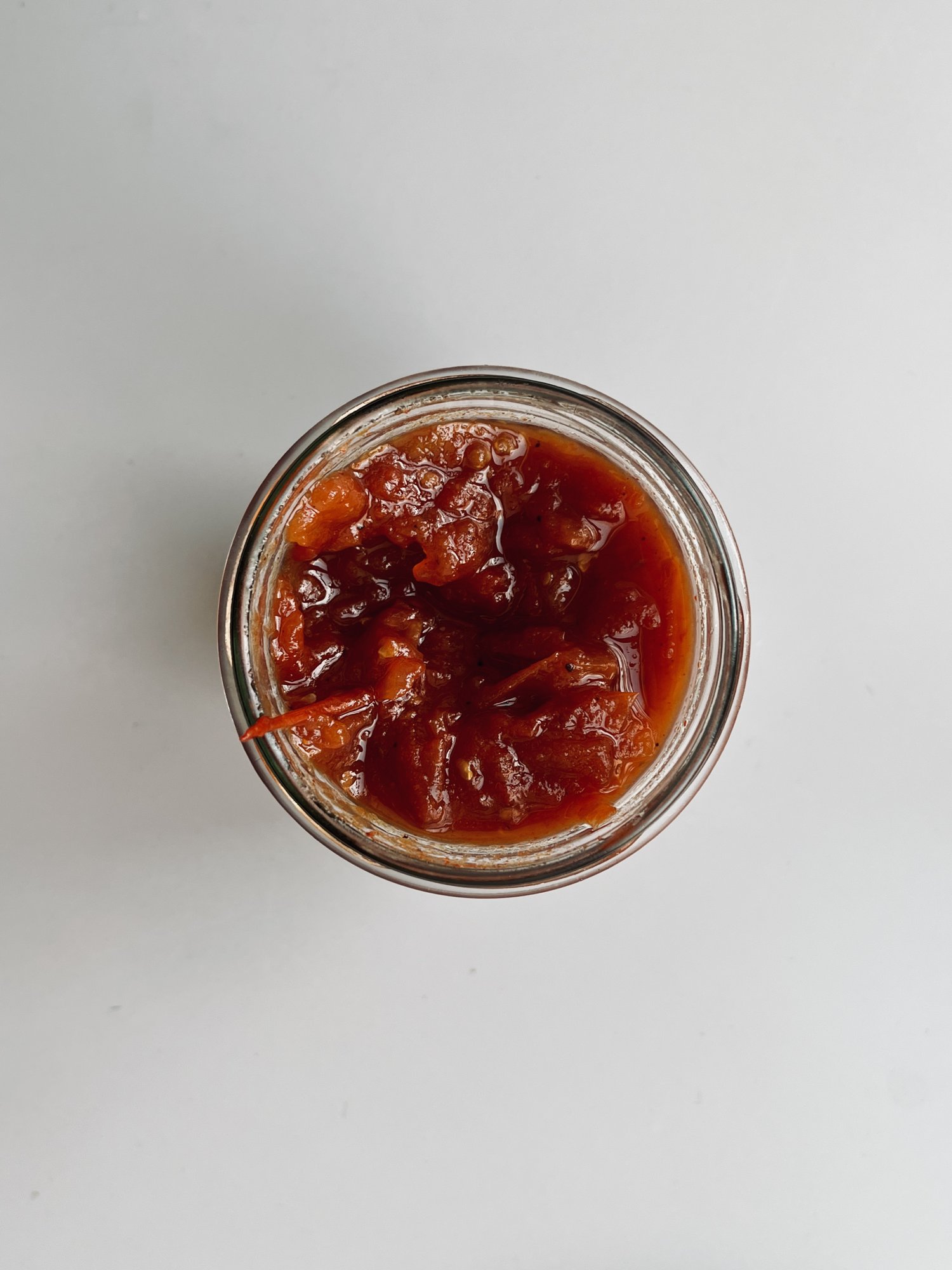 A small glass jar seen from above on a pale grey surface, filled with a thick, chunky red tomato jam flecked with dark spice.