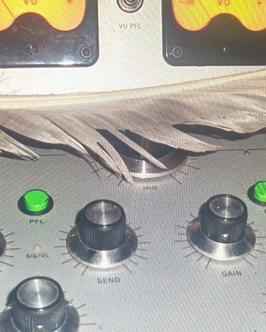 Close-up of an audio mixer: two orange-lit VU meters at the top, a lit green PFL button, and SEND and GAIN knobs, with dried leaves or pale plant fibre lying across the panel between them.